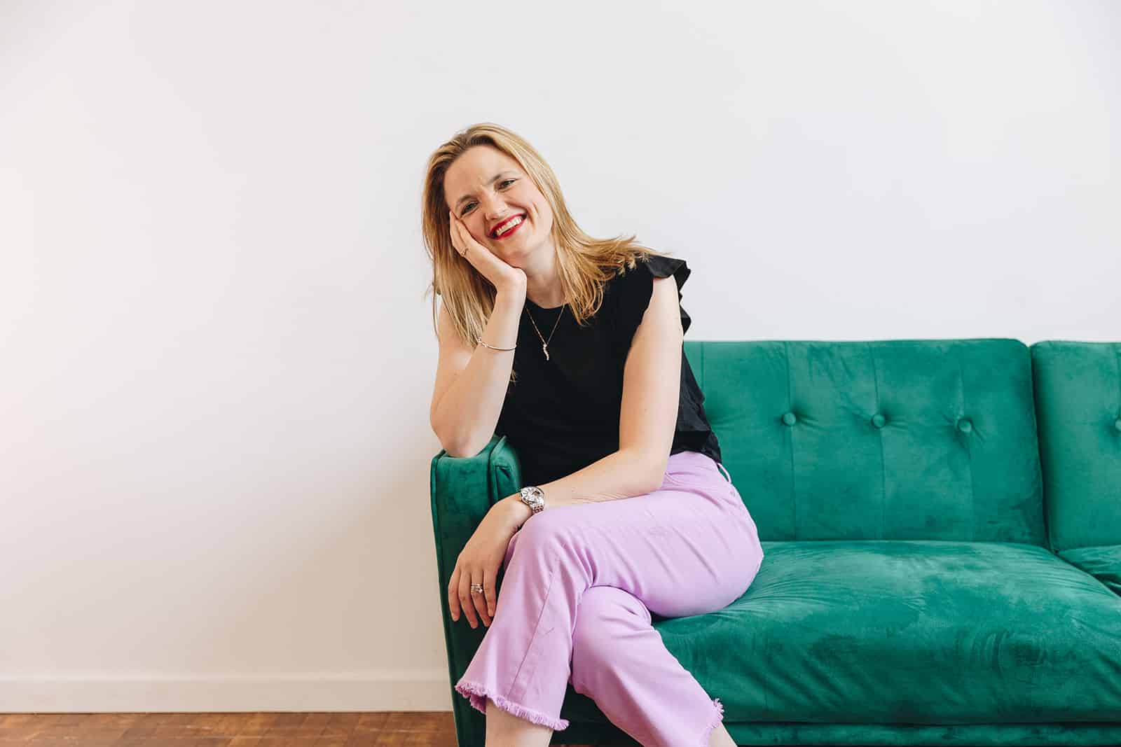 Building Momentum Brooke Greening, a sales coach, sitting on a green couch, smiling, and wearing a black top and purple pants.
